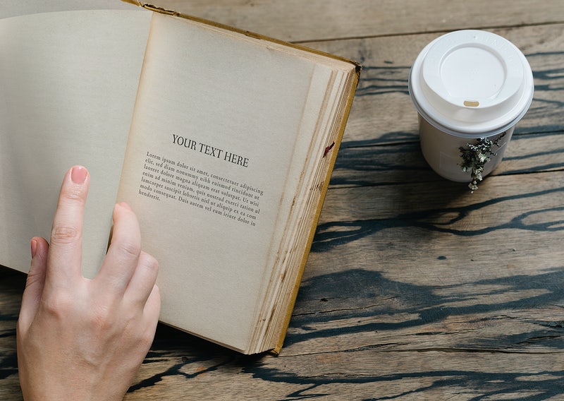 A person holding open an old book mockup with "your text here" placeholder text, next to a takeaway coffee cup on a wooden table.