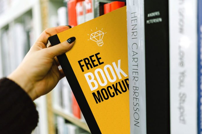 A hand holding a yellow book mockup with the words "free book mockup" and a lightbulb icon from a shelf in a library or bookstore.