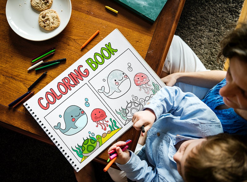 A child enjoys coloring in a sea-themed coloring book mockup with crayons, with freshly baked cookies nearby for a creative snack break.