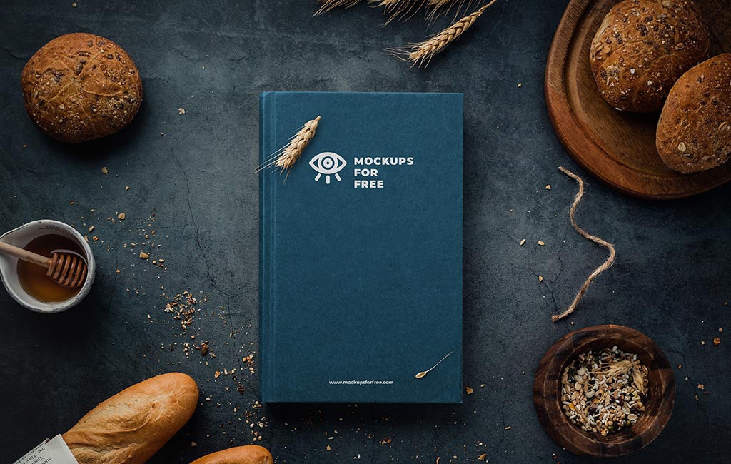 A minimalist bakery-themed flat lay featuring a dark blue book on table with a wheat illustration surrounded by artisan bread, grain ears, a bowl of seeds, and a jar of honey on a dark textured surface