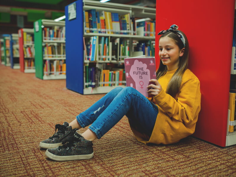 A young girl sitting on the floor of a library, leaning against a red bookshelf, engrossed in reading a book.