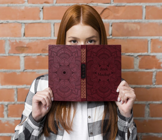 A woman holding a book with red hair peeking over an opened book with a decorative cover, against a brick wall backdrop. The book is labeled "book mockup" in the center.
