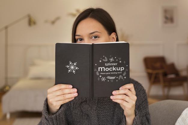 A woman in a cozy sweater holding up a black book with "winter mockup" written on the cover, partially covering her face.